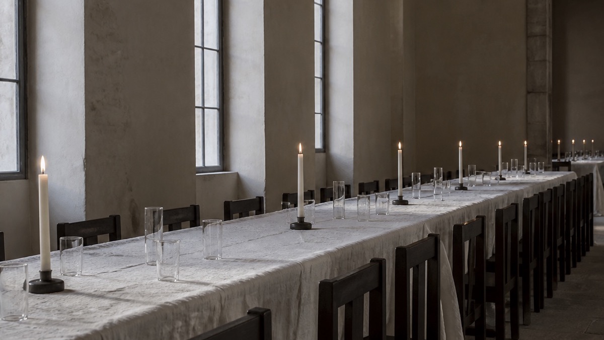 A long empty table under white linen with tall candles and clear glasses in a stone hall.